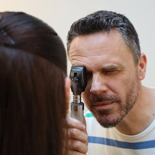 A male doctor in a stripy top using a Welch Allyn Coaxial ophthalmoscope on a female patient with long brown hair against a cream background.