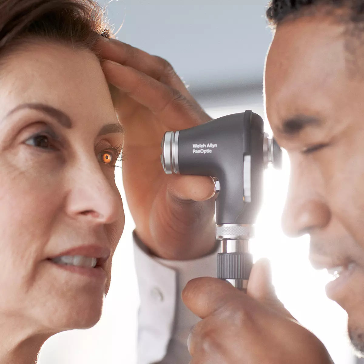 A male doctor is using the Welch Allyn PanOptic ophthalmoscope on a female patient. "Welch Allyn PanOptic" is clearly written on the side. They are in front of a window with light shining through. You can see the light on the ladies eye from where the ophthalmoscope is on.