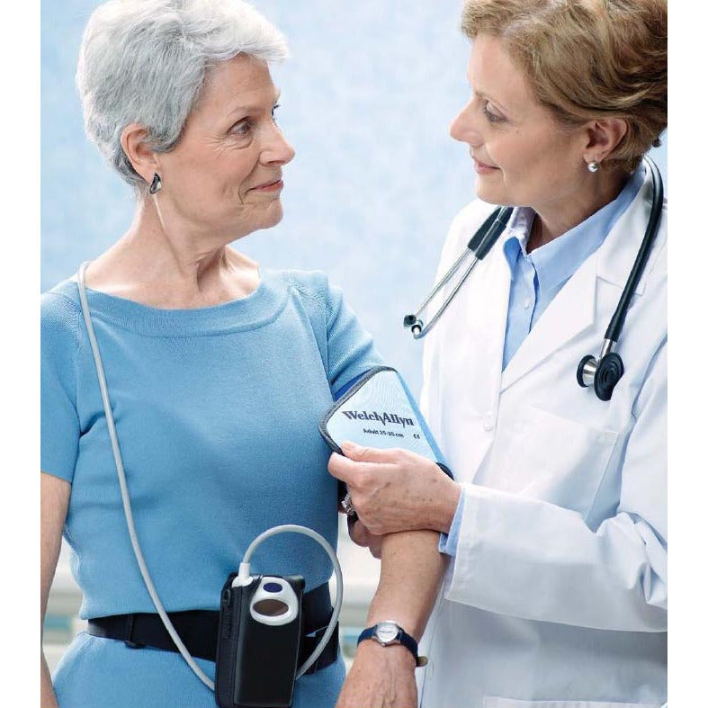 Welch Allyn black waist band around an old ladys waist with a Welch Allyn digital blood pressure monitor in the case and a tube leading to a blood pressure cuff a female doctor is putting on her arm. The paitent is wearing a light blue short sleeved jumper dress and the doctor is wearing a white lab coat and a blue shirt with a Welch Allyn stethoscope around her neclk. Pale blue background.