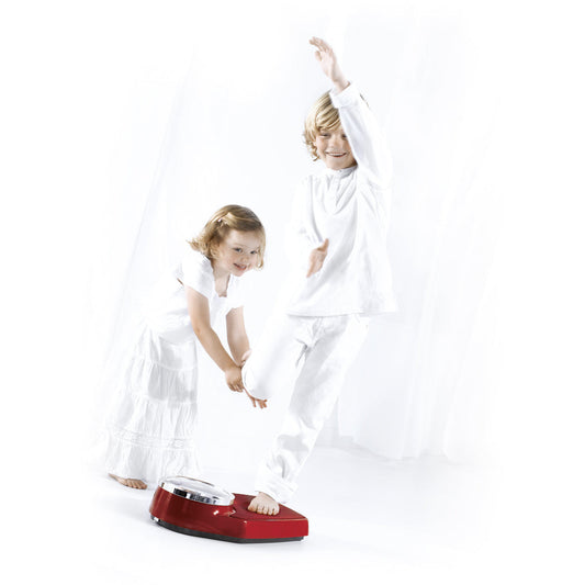 Tow kids both dressed in white playing in a white room with a boy standing with one foot on a set of burgundy seca 760 colorata non-medical flat scales and a little girl holding onto his other foot.