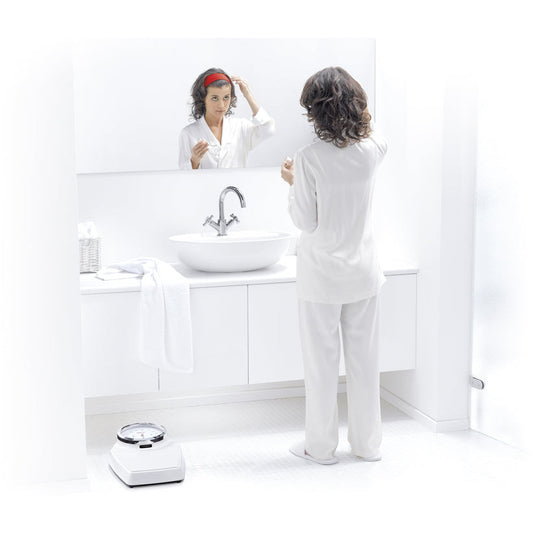 White seca 760 colorata non-medical flat scales in a white bathroom in front of a sink basin. A lady is looking into a long horizontal mirror whilst wearing a red headband and white clothes. The bathroom is white.